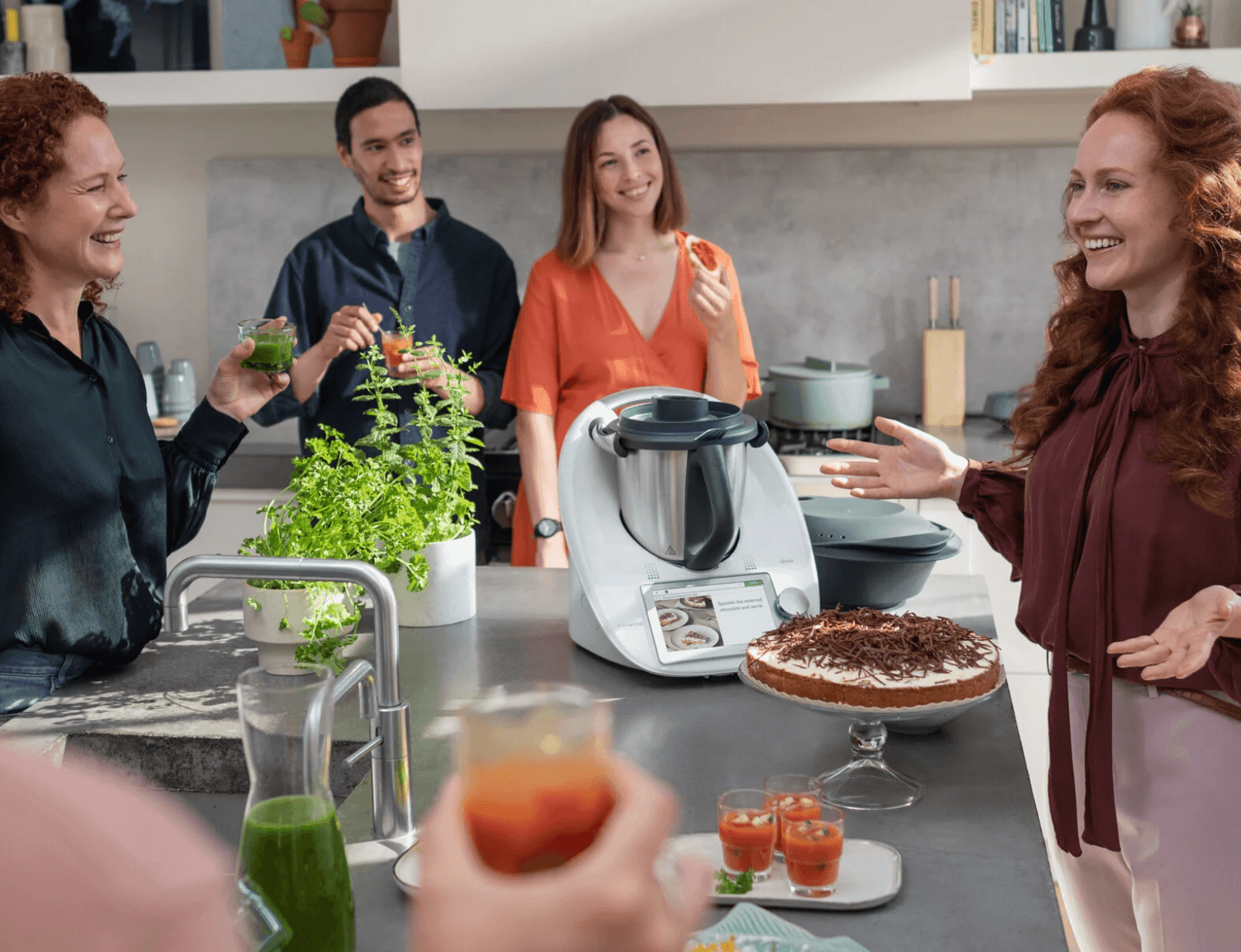 People gathered around a kitchen island with a cooking appliance and various drinks, discussing or enjoying refreshments.